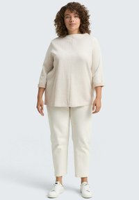 Donna con capelli ricci che indossa un maglione beige largo, pantaloni color crema e sneakers bianche, in piedi contro uno sfondo grigio chiaro.