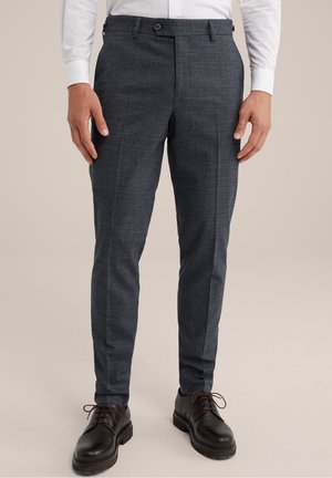 Mann trägt dunkelgraue karierte Slim-Fit-Anzughose, weißes Hemd und schwarze geschnürte Lederschuhe, steht vor schlichtem Hintergrund.