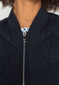 Quiltet marineblå bomberjakke med ribbet krave, sølv lynlås og broderet logo detalje. Glat stoftekstur.