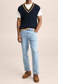 Marineblauer gerippter ärmelloser Pulli mit gelben Akzenten über einem weißen T-Shirt, kombiniert mit hellblauen geraden Jeans und braunen Schnürschuhen.