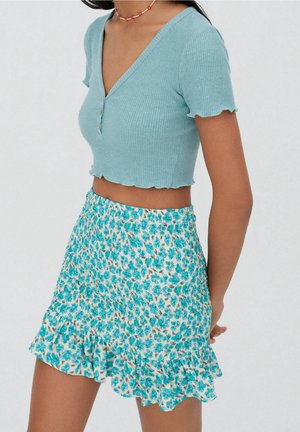 Femme portant un crop top côtelé à manches courtes bleu clair avec des boutons et une jupe froncée à motifs floraux blancs.