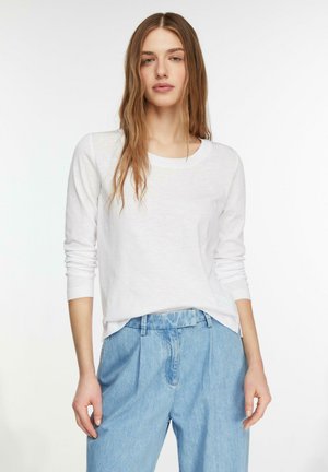 Woman with long hair wearing a white long-sleeve shirt and high-waisted light blue jeans, standing against a white background.