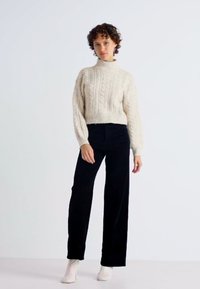 Pulover din lână cu model împletit, de culoare crem, cu guler înalt, asociat cu pantaloni largi din catifea neagră și ghete albe până la gleznă, pe un fond simplu.