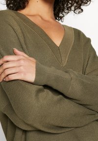 Pull en maille à col en V de couleur vert olive. Doté d'une texture côtelée, de poignets ajustés et d'une coupe décontractée. Le matériau semble doux et texturé.
