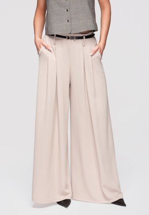 Kvinde iført beige bukser med vide ben og frontplissering, et sort bælte og en gråternet ærmeløs top, hænderne i lommerne.