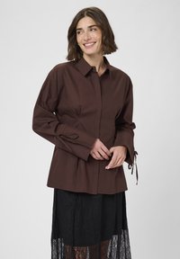 Brown button-up shirt with long sleeves, featuring tie accents at cuffs and a slightly fitted design. Worn over a black lace skirt.