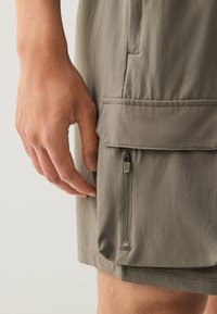 Shorts gris avec une poche zippée ; tissu léger et lisse ; ceinture plate ; main placée près de la poche pour le détail.