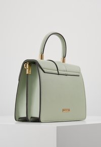 Zelená kožená kabelka se strukturovaným tvarem, zlatými kovovými doplňky a černým lemováním. Vybavena horním uchem a designem s klopou.