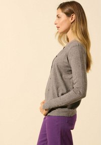 Cardigan gris avec encolure ronde, fermeture à boutons et manches longues, associé à un pantalon violet. Texture douce et design ajusté.