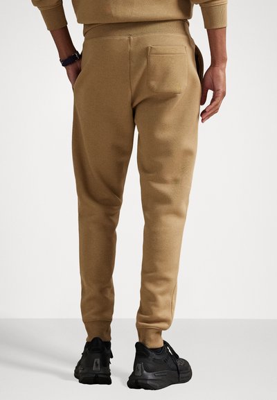 Pantaloni de trening bej cu croială ajustată, talie elastică și un buzunar pe spate. Purtați cu încălțăminte sport negre, cu o suprafață texturată.