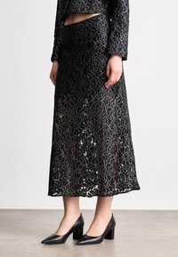 Juoda nėriniuota sijonas su gėlių raštu, pusiau permatoma konstrukcija ir tiesia siluetu, derinamas su žemakulniais juodais bateliais.