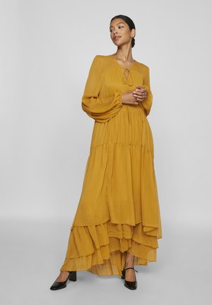 Maxi dress in mustard yellow, featuring tiered ruffles, long sleeves, a V-neck with ties, made of lightweight fabric, and paired with black heels.