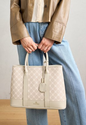 Personne portant une veste marron et un jean bleu clair tient un sac à main beige à motifs avec deux poignées et une étiquette de bagage.