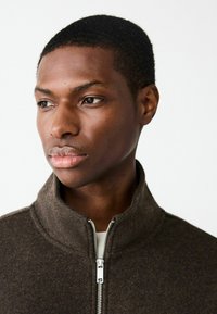 Hnedá zipsová bunda vyrobená z mäkkej, textúrovanej látky s vysokým golierom a strieborným zipsom. Vyznačuje sa priliehavým strihom a minimalistickým štýlom.