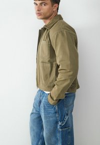 Olive green jacket with a collar and front pockets, paired with light blue baggy jeans featuring side pockets and a faded texture.