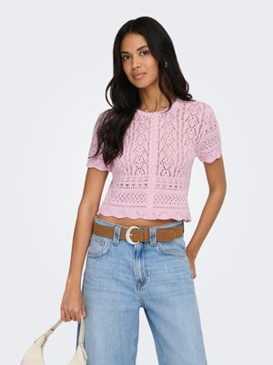 Mujer con cabello largo y oscuro que lleva una blusa rosa de encaje de manga corta, vaqueros de talle alto azul claro, cinturón marrón, sosteniendo un bolso blanco.