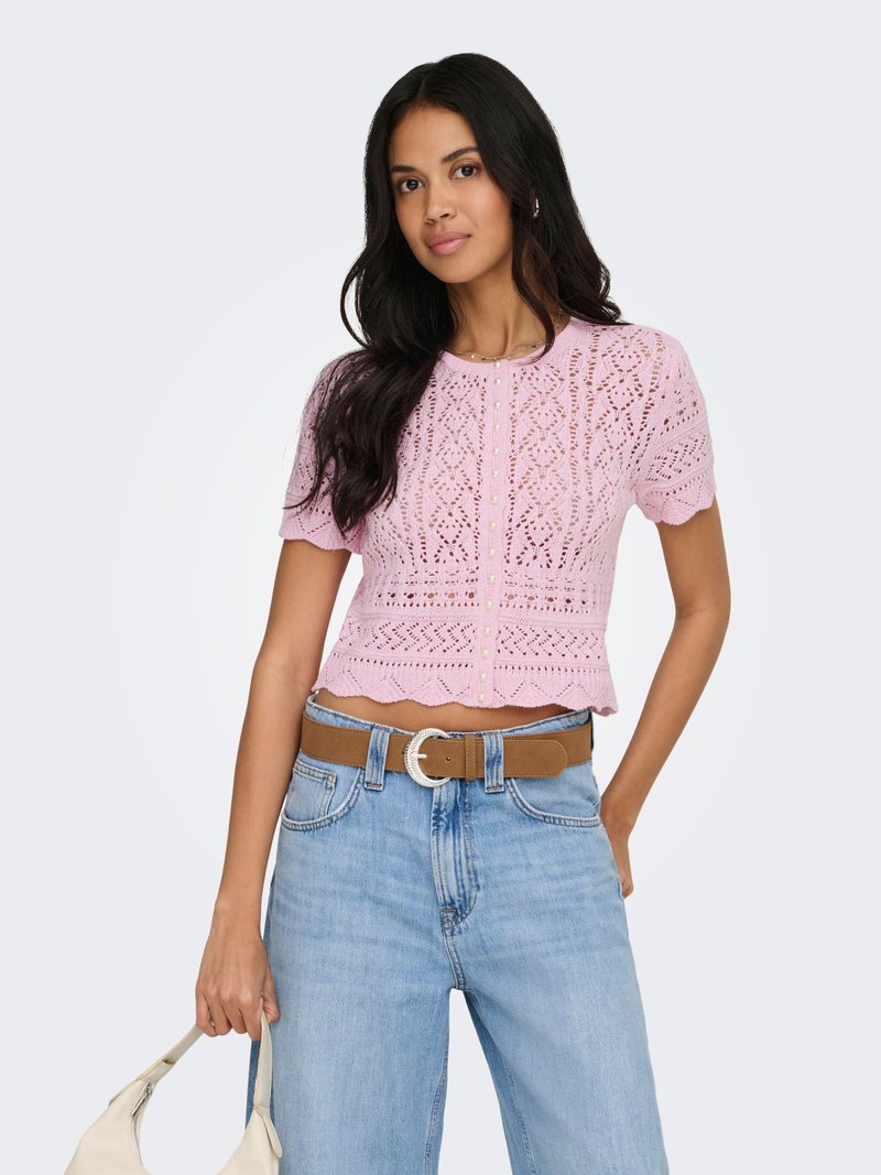 Femme aux longs cheveux foncés portant un haut court en dentelle rose à manches courtes, un jean taille haute bleu clair, une ceinture marron, tenant un sac à main blanc.