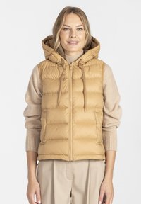 Beżowa kamizelka ocieplana z kapturem, z zapięciem na zamek błyskawiczny z przodu, regulowanymi sznurkami i kieszeniami bocznymi. Noszona na lekkim swetrze.