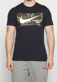 Camiseta negra de algodón, con un patrón de camuflaje y un gran logotipo de Nike en blanco en el frente. Mangas cortas y diseño de cuello redondo.
