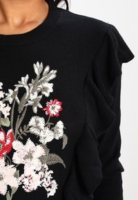 Pull en tricot noir avec broderie florale en blanc, rouge et vert. Présente des épaules froncées. Texture lisse.