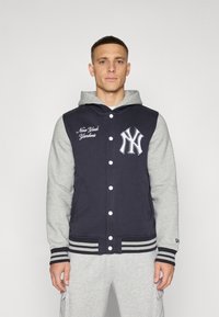 Chaqueta con capucha en color navy y gris, con cierre de botones en la parte delantera, logo de los New York Yankees y puños a rayas. Tejido suave con un diseño deportivo.