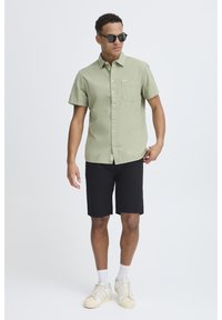 Lysegrønn kortermet skjorte med teksturert stoff, enkelt brystlomme, kombinert med svarte shorts og beige sneakers. Svarte solbriller blir brukt.