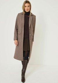 Brun uldblandingsfrakke med et struktureret design, klapkrave og to frontlommer. Sammen med en mørk striktrøje og brune knæhøje støvler.
