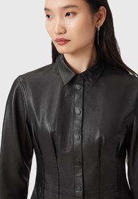 Abito camicia in pelle nera con chiusura a bottoni, colletto, maniche lunghe e cuciture strutturate per una vestibilità su misura.
