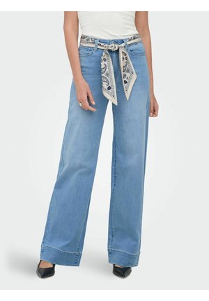 Žena nosi svetlomodré džínsové nohavice s širokými nohavicami, opasok z vzorovanej šatky, biely top a čierne špicaté topánky, stojí pred jednoduchým pozadím.