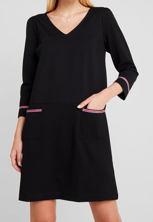 Schwarzes Kleid mit V-Ausschnitt, dreiviertel Ärmeln und zwei aufgesetzten Taschen mit rotem und weißem Besatz. Weicher Stoff und lockere Passform.