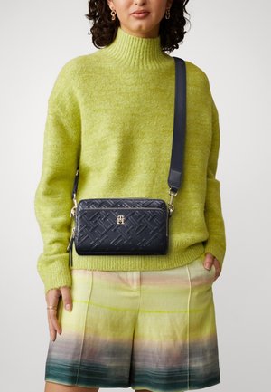 Navy leather crossbody bag with quilted pattern and gold logo, worn with a bright green sweater and multicolored shorts.