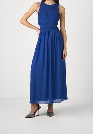 Femme portant une robe maxi bleu roi sans manches avec un corsage plissé et une taille découpée, associée à des escarpins pointus noirs et argentés.
