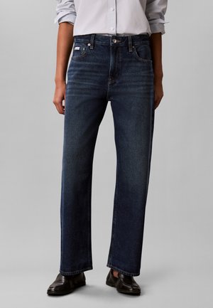 Person trägt dunkelblaue Jeans mit geradem Bein, weißes Hemd mit Knöpfen und hochgekrempelten Ärmeln sowie schwarze Schuhe und steht vor einem grauen Hintergrund.
