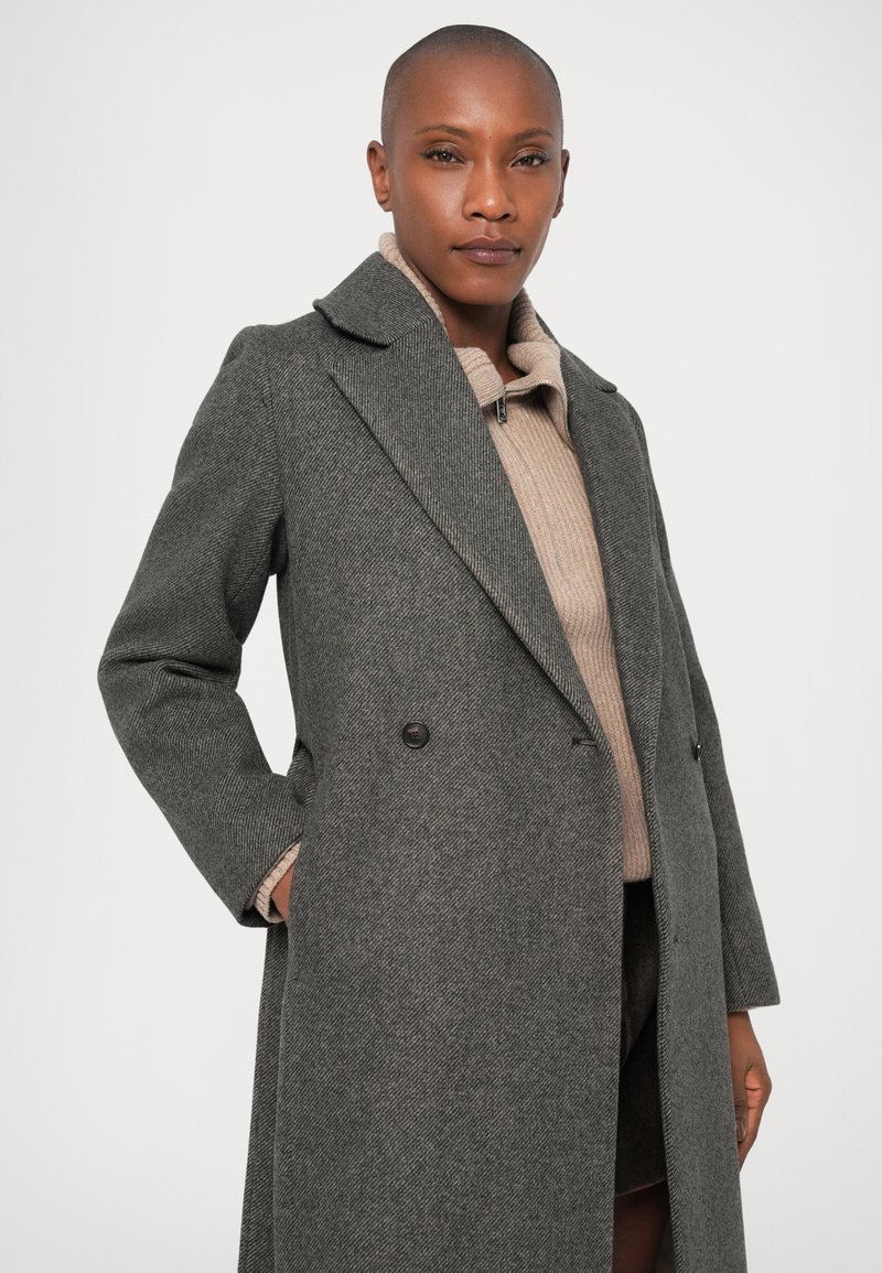 Grau melierter Wollmix-Mantel mit Fischgrätmuster, doppelt geknöpftem Design, breiten Revers und seitlichen Taschen. Getragen über einem beigen Strickoberteil.
