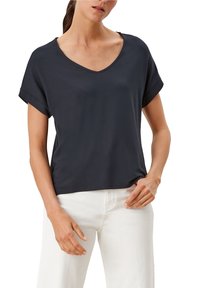 Eine Frau trägt ein dunkelgraues V-Ausschnitt-T-Shirt mit kurzen Ärmeln und weiße Hosen, wobei eine Hand auf ihrer Hüfte ruht und die andere entspannt an ihrer Seite hängt.