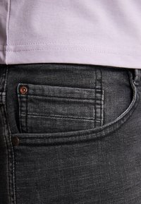 Jeans de mezclilla gris oscuro con un bolsillo trasero visible. La tela tiene una ligera textura y los jeans se combinan con una camiseta de color púrpura claro.