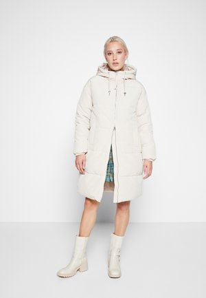 Parka rembourrée beige avec capuche, dotée d'une fermeture éclair à l'avant, de deux poches latérales et de finitions côtelées. Portée par-dessus une robe à carreaux bleu et vert.
