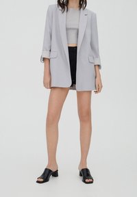 Blazer gris clair avec manches retroussées, associé à un crop top gris ajusté et un short noir, complété par des mules noires à bouts ouverts.