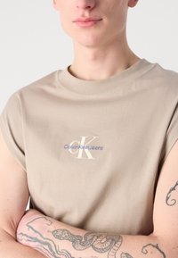 Beige trumpas rankovėmis marškinėliai, pagaminti iš minkštos medvilnės, su apvalia iškirpte. Puošia išsiuvinėtas „CK“ logo ir „Calvin Klein Jeans“ tekstas.