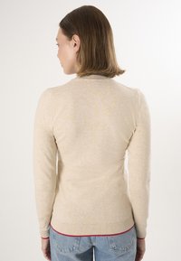 Beige gebreide trui met lange mouwen, geribbelde kraag en manchetten, en een rode accent aan de zoom. Glad textuur, getailleerd ontwerp.