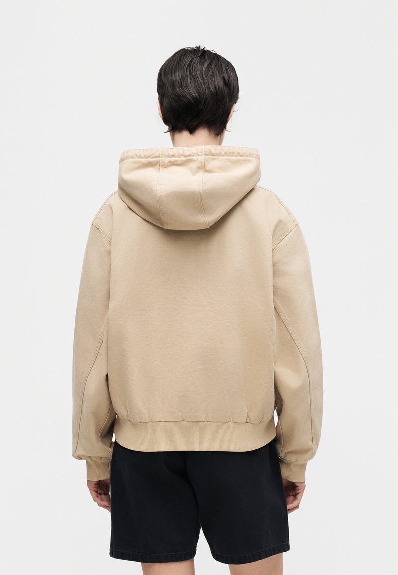 Personne de dos portant un sweat à capuche beige et un short noir sur un fond clair uni.