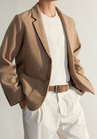 Blazer marrone in misto lana con revers a punta, chiusura a due bottoni e tasche applicate, indossato sopra una maglietta bianca a collo rotondo e una cintura beige.