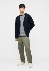 Homme portant un cardigan bleu marine, une chemise rayée noir et blanc, un pantalon vert olive et des baskets blanches, debout les mains derrière le dos.