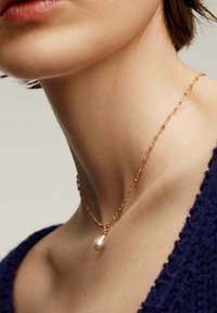 Collar de cadena de oro con pequeños acentos rosas y un único colgante de perla en forma de lágrima, usado en un cuello desnudo.