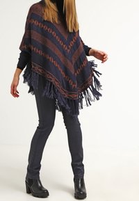 Kvinna iklädd en marinblå och brun mönstrad fransad poncho, svarta åtsittande byxor och svarta klackade ankelstövlar, stående mot en vit bakgrund.