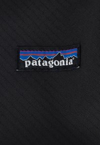 Černá tkanina s texturovaným povrchem obsahuje přišitý obdélníkový nášivku s logem Patagonia v bílé, modré a fialové barvě.