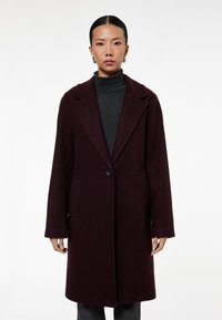 Bordeaux uldfrakke med enkelt knaplukning, spidse lapels og sidelommer; parret med en grå turtleneck. Glat tekstur.