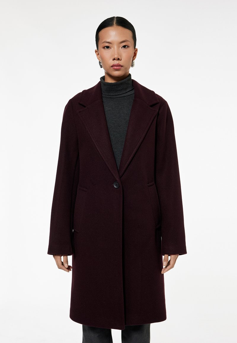 Bordeaux uldfrakke med enkelt knaplukning, spidse lapels og sidelommer; parret med en grå turtleneck. Glat tekstur.