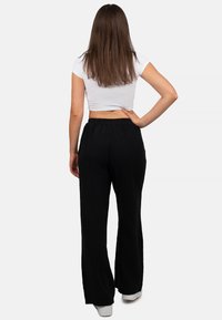 Zwarte, wijde pantalon in een gestructureerde stof met een elastische tailleband. G gedragen met een korte witte top, van achteren gezien, met witte schoenen.