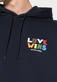 Sudadera de color azul marino con textura acanalada. Presenta el texto bordado multicolor "LOVE WINS" y el nombre de la marca "SCOTCH & SODA" debajo.